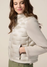 Jachetă puffer de culoare bej deschis, cu o textură netedă, având un guler înalt și buzunare cu fermoar, asortată cu un top lung cu mâneci de culoare gri deschis în contrast.