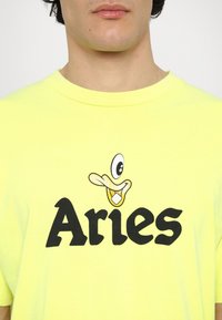 T-shirt di cotone giallo con grafica di un'anatra cartone animato sopra la parola "Ariete" in grassetto nero. Maniche corte e scollatura tonda.