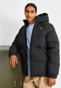 Schwarze Bomberjacke mit Reißverschluss vorne, Kapuze und elastischen Bündchen. Mit einem Logo auf der Brust und gestepptem Design. Wird über einem hellblauen Hemd getragen.