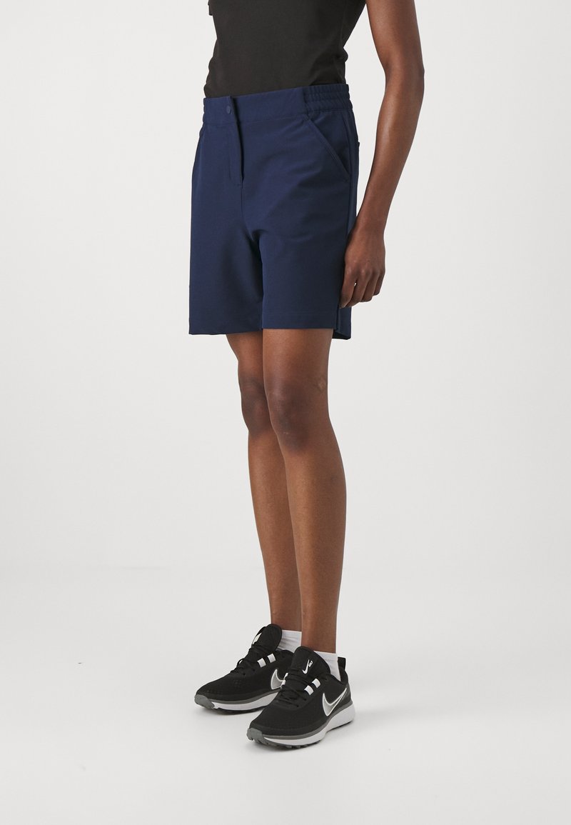 Shorts bleu marine en tissu lisse et extensible, dotés de poches latérales et d'un devant plat. Associés à des chaussures de sport noires et des chaussettes blanches.