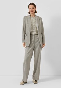 Vrouw in een lichtgrijs pak met wijde broekspijpen, een bijpassende blazer, een zilver met pailletten bedekte top en metallic blokhakken schoenen.