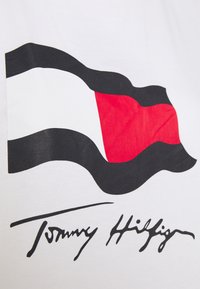 Camiseta blanca con un diseño de bandera impreso en rojo, blanco y negro, con la firma "Tommy Hilfiger" en negro debajo del gráfico.