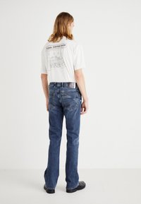 Camisa branca com um gráfico de café nas costas, combinada com jeans de ganga azul. Os jeans possuem um design de perna reta e costuras visíveis.