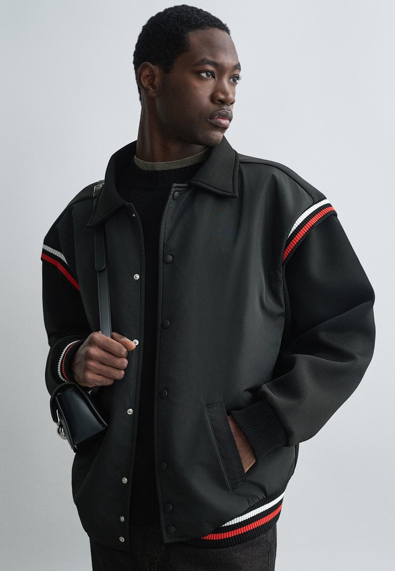 Mustam bomber-jakk nööbidest, kontrastsete punaste ja valgete triibuliste aktsentidega varrukatel ning soonikkudega mansettidel. Tekstuuriga kangas, ees taskud.