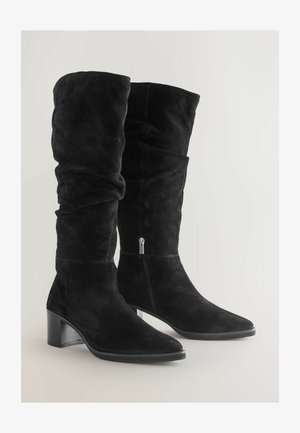 Czarne zamszowe kozaki do kolan o luźnym kroju, kwadratowym nosku i słupkowym obcasie. Posiadają srebrny zamek błyskawiczny z boku.