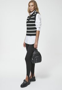 Czarny i biały paskowany sweter bez rękawów nałożony na białą koszulę z guzikami, w połączeniu z czarnymi leginsami i butami, trzymając mały czarny plecak.