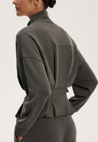 Siva, dolgi rokav jakna iz mehke tkanine; ima visok ovratnik, prilegajoč kroj in guban detajl na hrbtu s pasom.