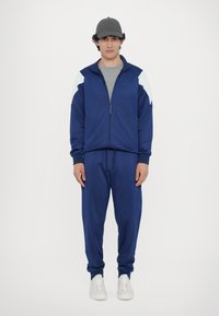 Tuta sportiva blu navy con giacca con zip e pantaloni con vita elastica, caratterizzati da accenti azzurri sulle spalle e sneakers bianche.