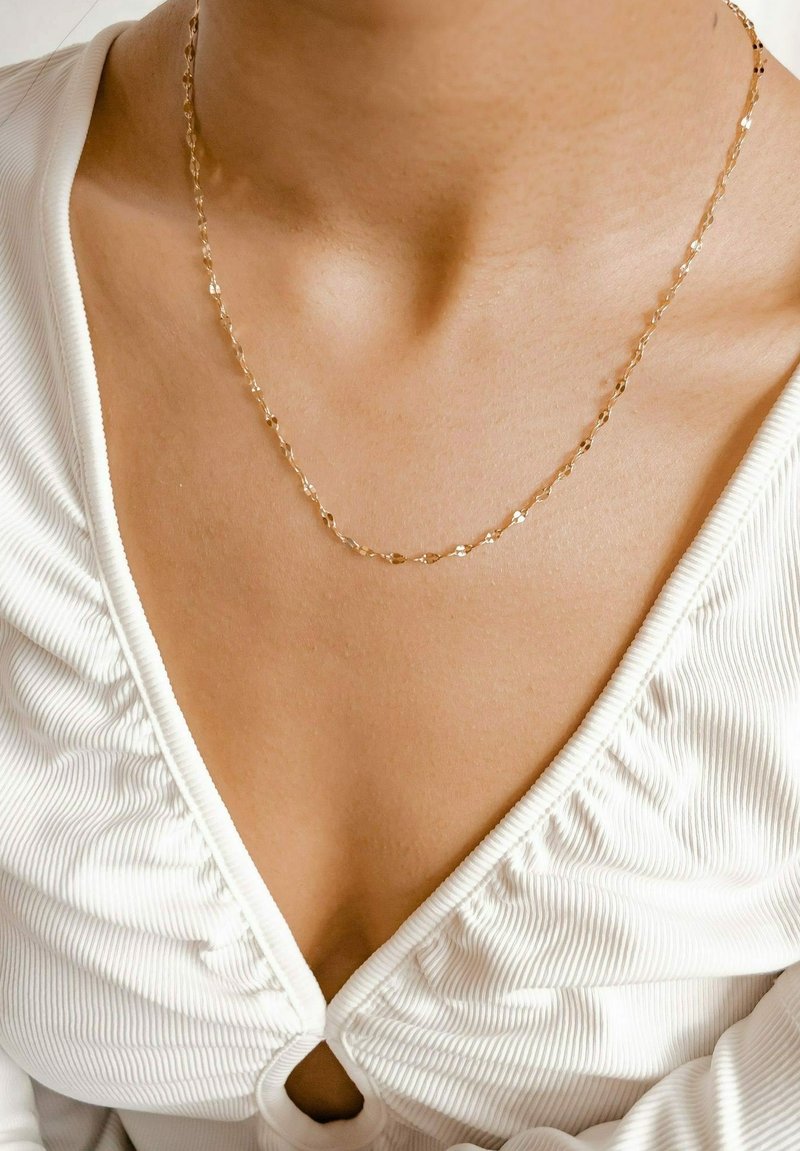 Collar de tono dorado con eslabones alargados y facetados, reposando sobre una superficie de piel suave, combinado con una blusa blanca texturizada.