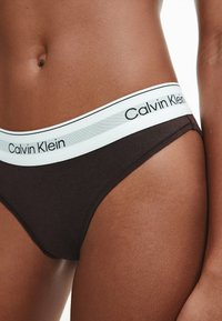 Tanga de algodón negro con una cinturilla blanca y gris que presenta el logo "Calvin Klein". Vista en primer plano que resalta el material y los detalles del diseño.