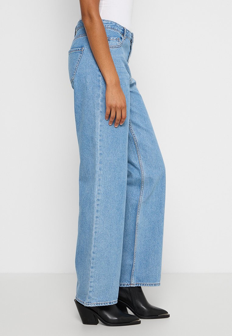 Jasnoniebieskie spodnie o szerokich nogawkach, z wysokim stanem, w klasyczny wzór z pięcioma kieszeniami i prostymi nogawkami, zestawione z czarnymi botkami na obcasie.