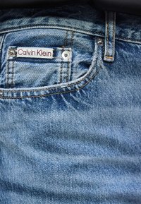 Nahaufnahme der Tasche einer blauen Jeans mit einem Calvin Klein Etikett, das oberhalb der kleinen Münztasche aufgenäht ist, und sichtbaren Metallnieten.