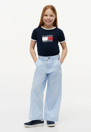 Niña sonriente de pie con las manos en los bolsillos, vistiendo una camiseta azul marino de Tommy Hilfiger, pantalones de pierna ancha en azul claro y zapatillas deportivas azul marino sobre un fondo blanco.