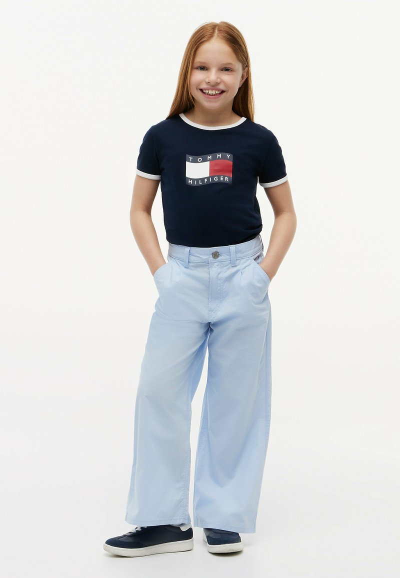 Ragazza sorridente in piedi con le mani nelle tasche, indossa una t-shirt Tommy Hilfiger blu navy, pantaloni larghi color azzurro chiaro e sneakers blu navy su sfondo bianco.