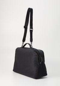 Sac en nylon noir de forme rectangulaire, avec des poignées doubles, une sangle d'épaule réglable et une fermeture éclair. Texture lisse sans motifs visibles.