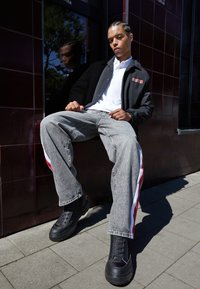 Chaqueta negra con capucha y logo, camiseta polo blanca, jeans grises con franjas laterales rojas y blancas, y zapatillas negras gruesas.