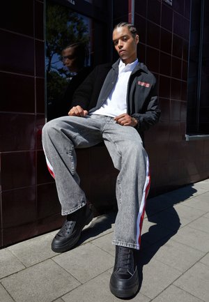 Chaqueta negra con capucha y logo, camiseta polo blanca, jeans grises con franjas laterales rojas y blancas, y zapatillas negras gruesas.