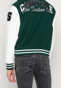 Giacca varsity verde e bianca con la scritta "COUTURE" e "The Couture Club". Presenta polsini e vita a coste a righe.