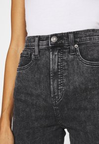 Vaqueros negros de cintura alta hechos de denim, con cierre frontal de botón, dos bolsillos y un ligero desgaste con detalles de costura visibles.