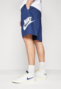 Granatowe spodenki Nike wykonane z lekkiego materiału, z dużym białym logo Swoosh i bocznymi kieszeniami. Zestawione z białymi sneakersami i skarpetkami.
