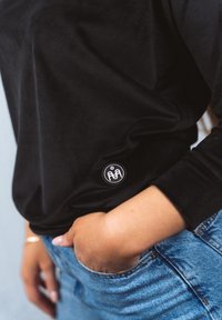 Musta pitkähihainen paita, jossa on teksturoitu kangas ja pyöreä merkki alaosassa, yhdistetty vaaleansinisiin farkkuihin.