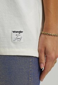 Hvid T-shirt med et stofmærke, der står "Wrangler with love, Lainey," parret med blå denim jeans med synlige sømme og talje.