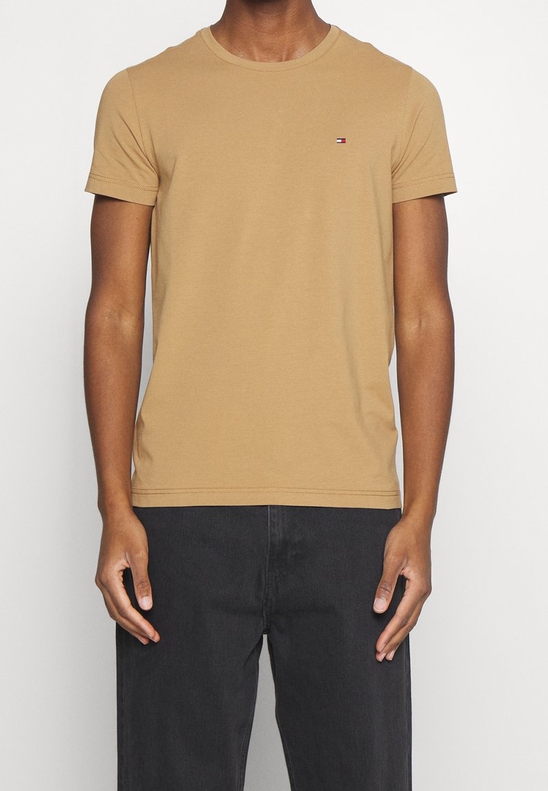 T-shirt en coton beige à manches courtes avec un col rond, présentant un petit logo brodé sur la poitrine et une silhouette ajustée.