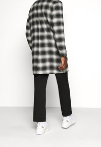 Cappotto lungo a scacchi bianco e nero con una vestibilità rilassata, abbinato a pantaloni neri e sneakers bianche. Il tessuto sembra strutturato e stratificato.