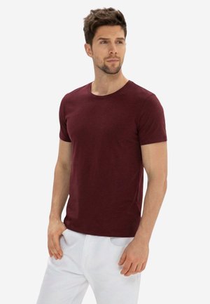 Junger Mann mit kurzen braunen Haaren, der ein eng anliegendes, kastanienbraunes kurzärmeliges T-Shirt und weiße Hose trägt, steht mit den Händen in den Taschen.