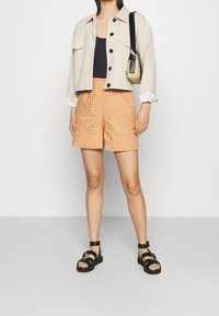 Veste beige cropped avec fermeture à boutons, haut marine et shorts en eyelet orange. Des sandales noires à lanières complètent la tenue.