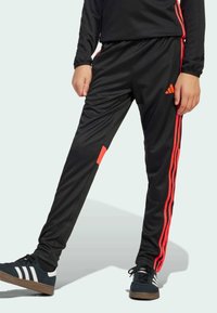 Svarta träningsbyxor med oranga ränder, tillverkade av slätt tyg. Har en orange flik på sidan och en Adidas-logotyp vid höfterna.