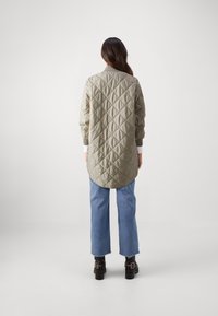 Chaqueta acolchada en verde claro con un dobladillo trasero más largo, puños elásticos y un corte relajado, combinada con jeans de pierna ancha azules y botas negras.