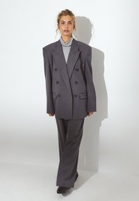 Blazer grigio doppiopetto con spalle oversize, abbinato a pantaloni larghi coordinati. Collo alto grigio testurizzato sotto.