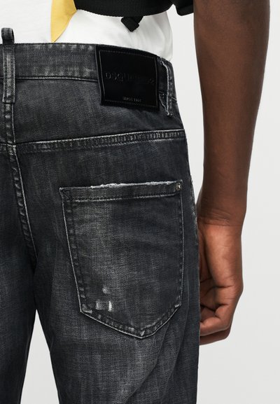 Οπίσθια όψη ενός ατόμου που φορά ξεθωριασμένο μαύρο τζιν με μαύρη ετικέτα DSQUARED2 πάνω από την πίσω τσέπη και ένα λευκό πουκάμισο μέσα στην μέση.