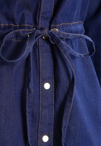 Tessuto denim blu scuro con cintura in vita annodata, occhielli dorati e bottoni a pressione bianchi lungo la parte anteriore e i polsini.