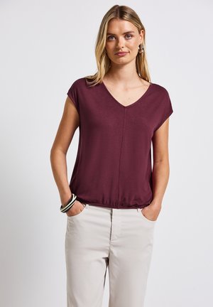 Top burdeos de manga corta con escote en V y dobladillo fruncido. Tela suave con un corte relajado, combinado con pantalones de color claro.