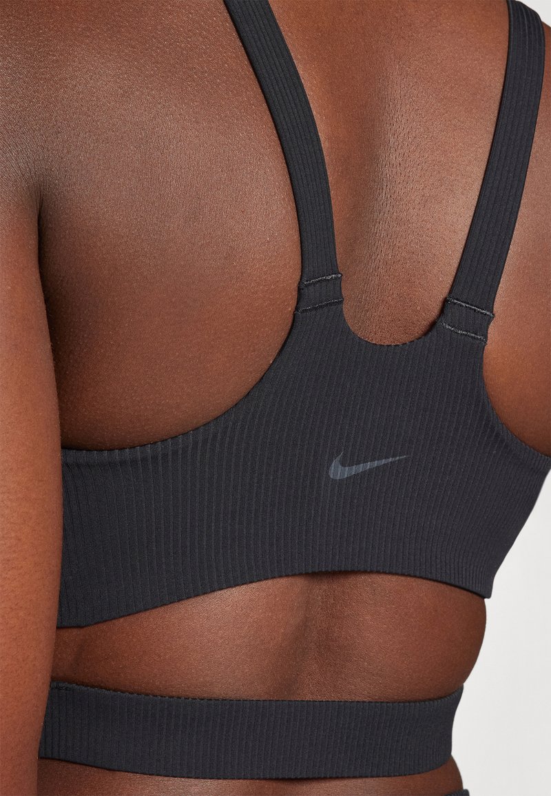 Sujetador deportivo negro acanalado con tirantes anchos y espalda de copa. Cuenta con un sutil logo de Nike en gris. Textura suave con un diseño ajustado.