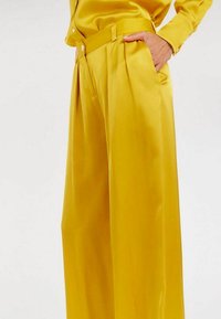 Pantalon large en satin jaune avec taille haute, devant plissé et poches latérales. Texture lisse et brillante, avec une coupe ajustée.