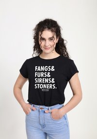 Mladá žena s kudrnatými vlasy, ruce v kapsách, oblečená v černém tričku s bílým textem "FANGS& FURS& SIRENS& STONERS." a světle modrých džínách.