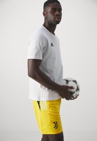 Camiseta de algodón blanca con mangas cortas y logo negro de Adidas; pantalones cortos amarillos con logo negro de Juventus y detalles en negro; sosteniendo un balón de fútbol en blanco y negro.