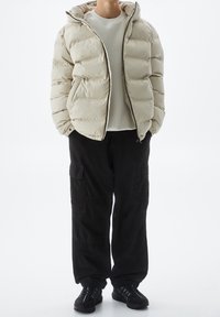 Lichtbeige gewatteerde jas met capuchon, voorzien van doorgestikte delen en een voorrits, gedragen over een grijs shirt en zwarte cargobroek. Zwarte schoenen.