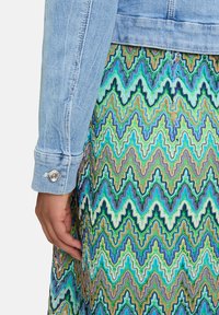 Manche de veste en denim bleu clair avec un bouton en argent, associée à une robe à motif zigzag multicolore dans des teintes de vert, bleu et marron.