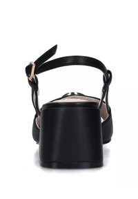 Sandalo nero con tacco largo e solido e cinturino alla caviglia. Presenta una texture in pelle liscia e un design minimalista. Vista posteriore.