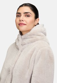 Manteau à capuche en fausse fourrure beige clair, présentant une surface texturée et une coupe décontractée. Capuche et fermeture avant visibles.