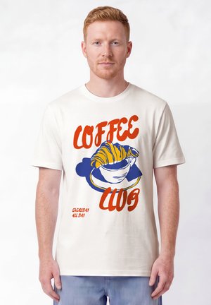 Homme aux cheveux roux porte un t-shirt blanc avec un texte orange "Coffee Club" et une illustration d'une tasse de café avec un croissant.