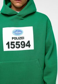 Groene hoodie van zachte stof met een voorkant zak, met een groot wit label waarop "POLIZEI 15594" in het zwart is gedrukt.