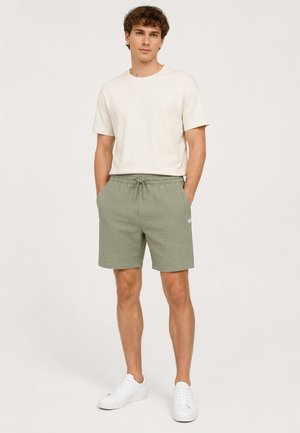 Ein Mann steht nach vorne gerichtet und trägt ein beigefarbenes T-Shirt, salbeigrüne Shorts mit Kordelzug und weiße Sneaker vor einem schlichten, hellen Hintergrund.