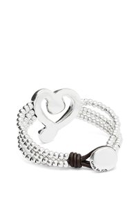 Centro de mesa en forma de corazón plateado sobre una banda de cuentas metálicas entrelazadas, con un acento de cuero marrón y detalles metálicos. Marcado "Uno de 50."