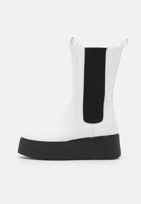 Bota branca até ao tornozelo com painéis laterais elásticos pretos e uma sola grossa preta. Couro liso com detalhe de costura e alças para puxar.