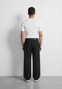 Άνδρας που στέκεται και κοιτάζει έναν λευκό τοίχο, φοράει λευκό πουκάμισο με κοντά μανίκια, μαύρο παντελόνι φαρδιάς γραμμής, μαύρη ζώνη και λευκά αθλητικά παπούτσια.